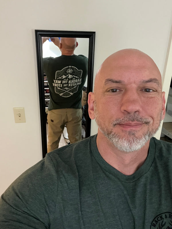 Man taking a mirror selfie wearing a Rack & River Outdoors tee, with the back graphic ‘Harder the Way, Greater the Story’ visible in the reflection.