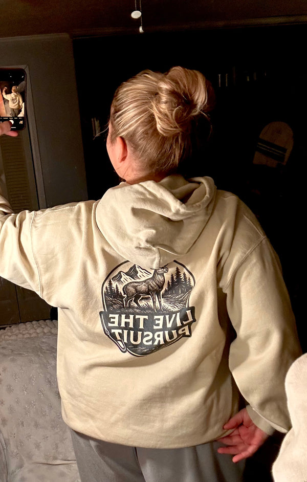 Woman wearing a Live the Pursuit deer graphic hoodie from Rack & River Outdoors, photographed from the back indoors.