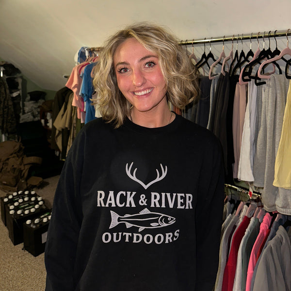 Woman wearing Rack & River Outdoors black crewneck with deer antlers and fish logo, standing in a room with hanging clothes. Christian outdoor apparel brand for hunters, anglers, and faith-driven lifestyles.