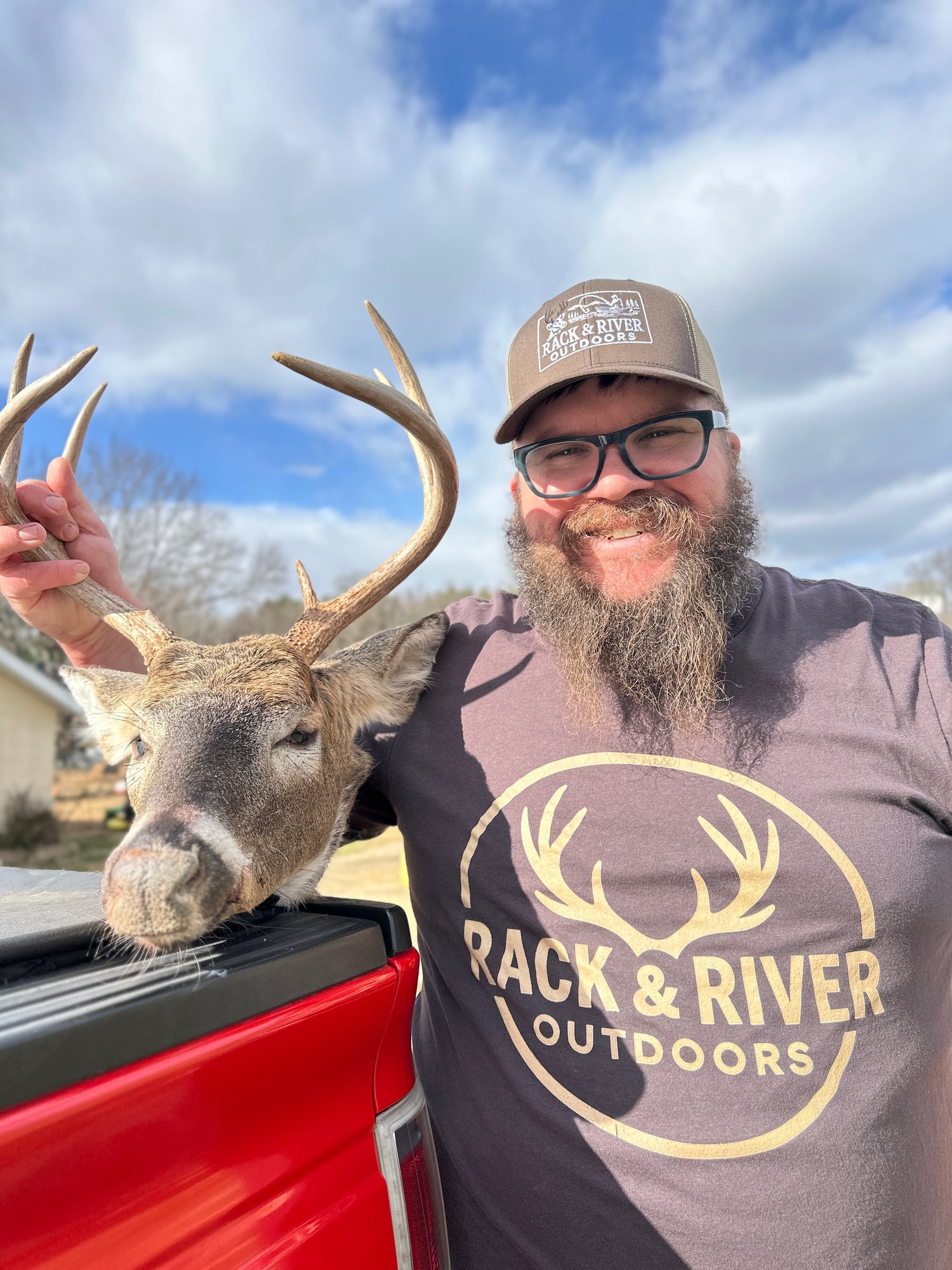 Man wearing a Rack & River Outdoors t-shirt and hat holding a harvested buck, standing beside a red truck on a sunny day. Christian outdoor apparel for hunters and nature lovers.